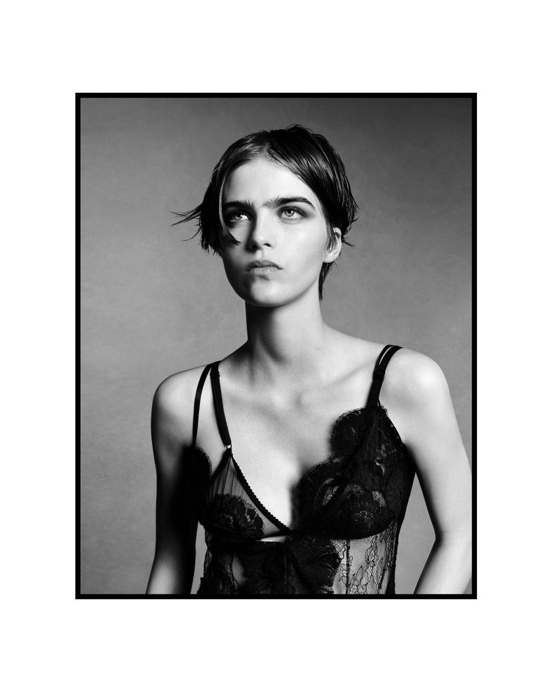 Monochrome portrait of a woman with a short haircut wearing a lace lingerie top against a neutral gray backdrop in a studio setting.