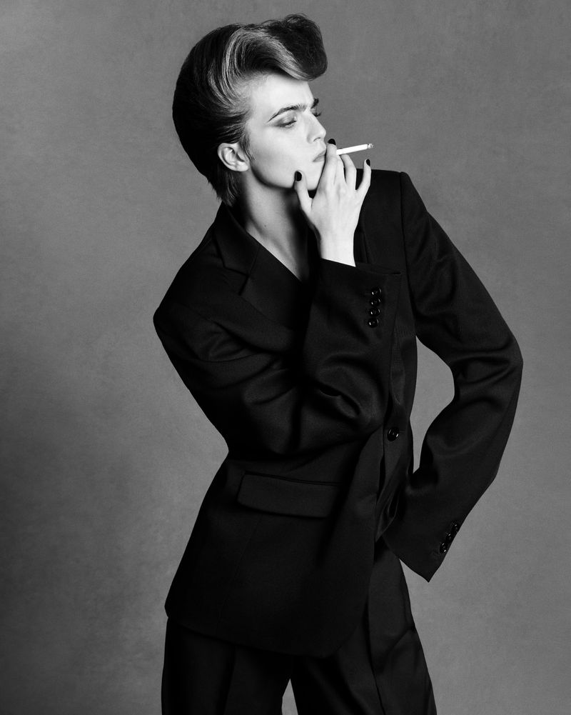 Black and white studio portrait of a woman in a suit smoking a cigarette, showcasing her stylized hair and fashionable attire.