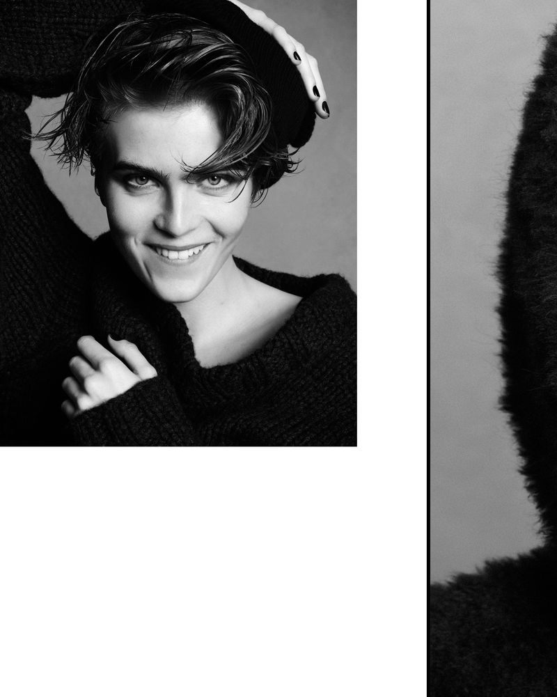 Black and white close-up portrait of a young adult smiling, wearing a dark sweater with a hand near the head, and a neutral background.