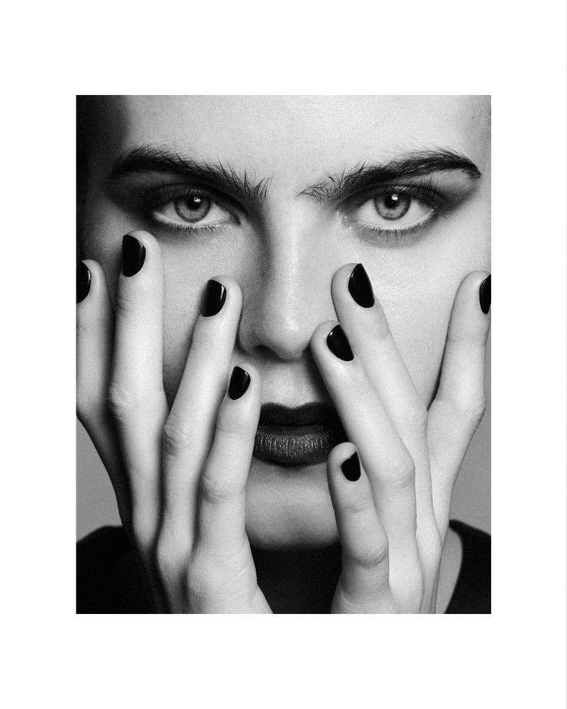 Monochrome portrait of a woman with hands near her face, showcasing dark lipstick and nail polish against a neutral gray backdrop.