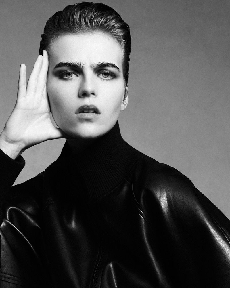 Black and white portrait of a woman with slicked-back hair wearing a black turtleneck and leather jacket, posing against a grey backdrop.