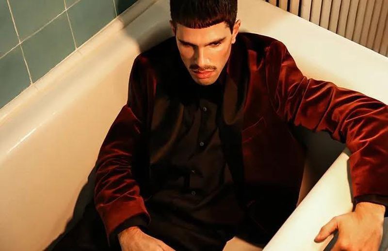 Portrait of an adult male with dark hair and a mustache, wearing a velvet jacket and a black shirt, sitting in a bathtub.