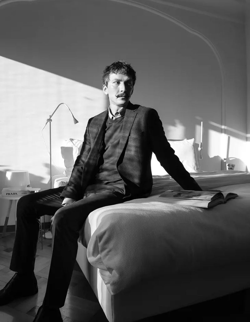 Black and white portrait of a moustached man in a suit sitting on a bed in bright sunlight. Book and lamp are visible.
