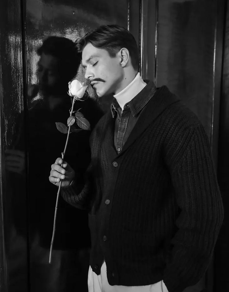 Black and white portrait of a man with a mustache wearing a cardigan, smelling a white rose. The photo has a vintage look.