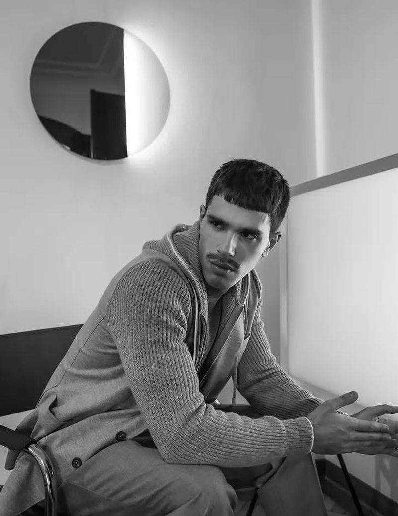 Black and white portrait of an adult male with a moustache sitting indoors, wearing a hooded sweater with a round light fixture in the background.