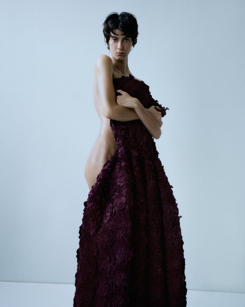 Portrait of a woman with dark hair holding a deep burgundy, textured textile against a light background in a fashion photography style.