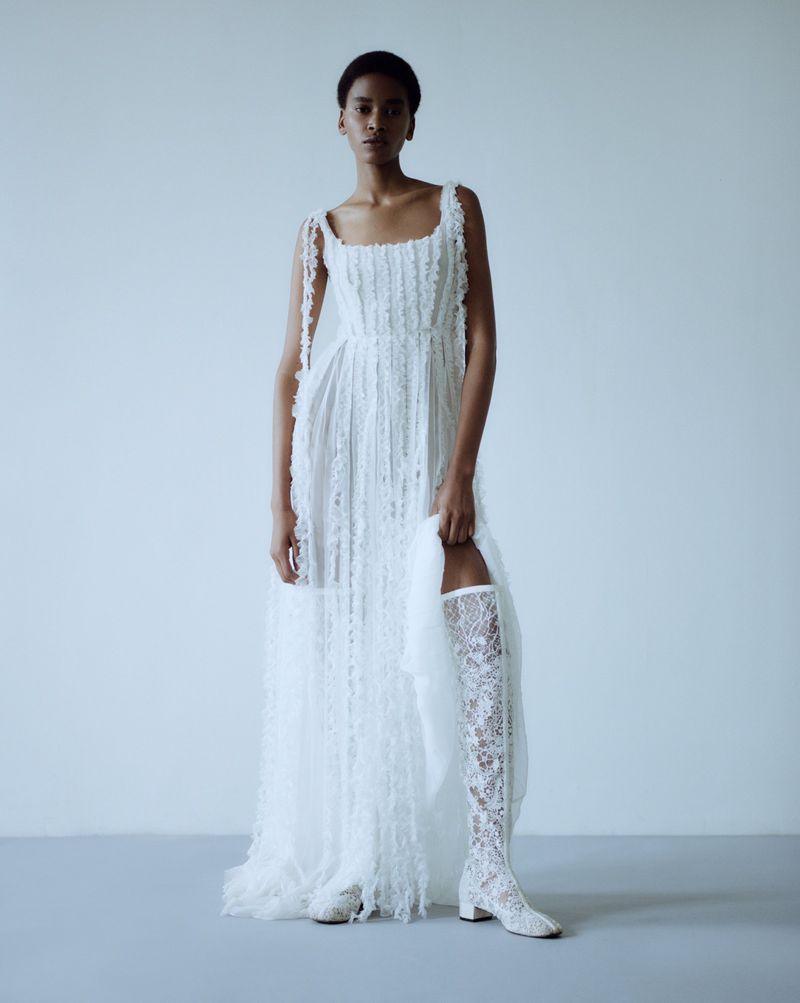 Woman in a long, white, lace dress and lace boots poses against a light blue background in a studio fashion shot.
