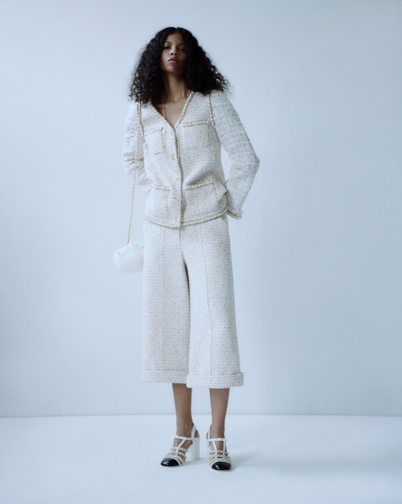 Full-length shot of a black female model in a white tweed jacket and culottes, with a small bag, posing against a white backdrop.