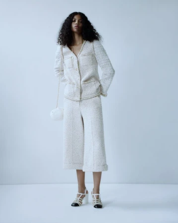 Full-length shot of a black female model in a white tweed jacket and culottes, with a small bag, posing against a white backdrop.
