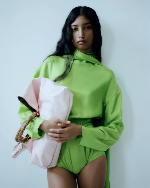Model wears a green satin top and shorts with a pink satchel. Studio shot with a neutral background, fashionable and modern aesthetic.