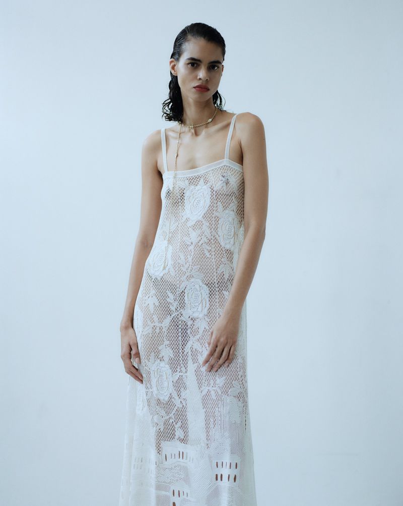 Model in a sheer white lace slip dress standing against a plain pale studio background for editorial shoot.