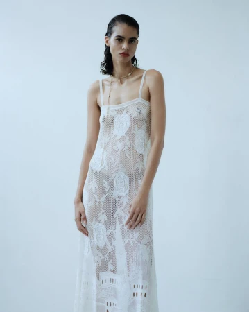 Model in a sheer white lace slip dress standing against a plain pale studio background for editorial shoot.