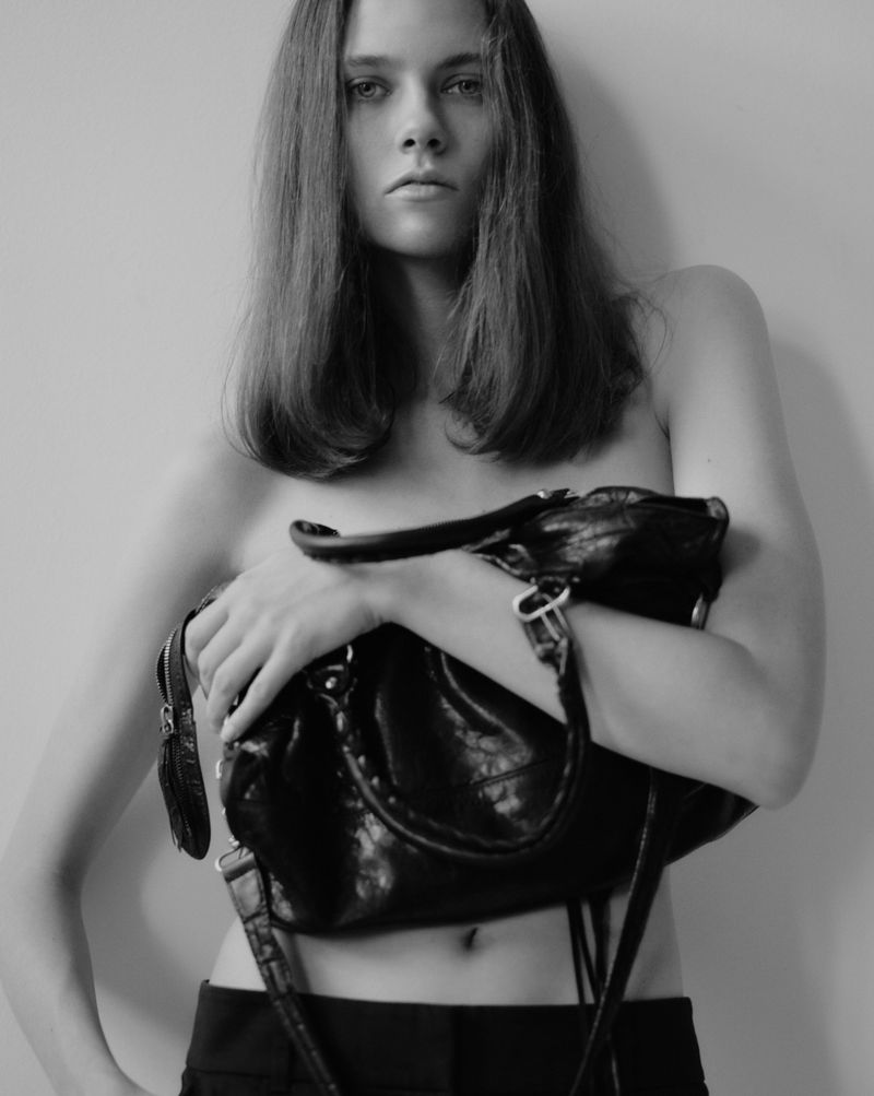 Black-and-white portrait of a topless young woman with dark hair, holding a large black purse in front of her torso.