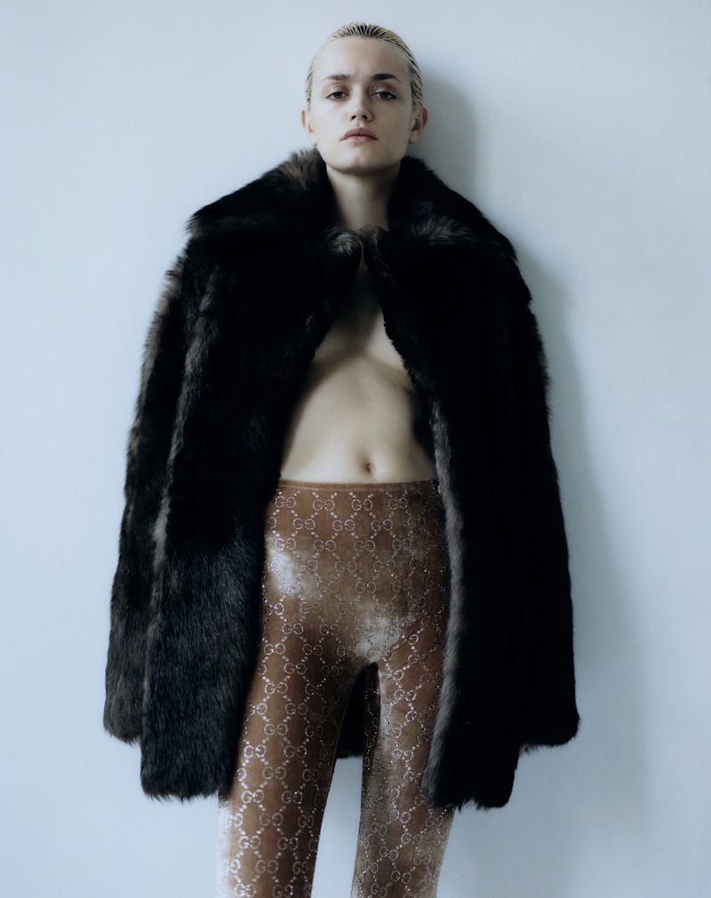A woman in fur coat and patterned leggings poses against a white wall in a studio portrait, fashion shoot, beauty and style.