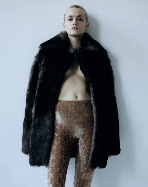 A woman in fur coat and patterned leggings poses against a white wall in a studio portrait, fashion shoot, beauty and style.