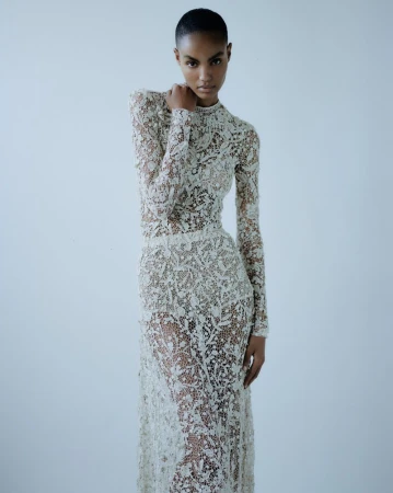A slender African-American fashion model poses in a studio, wearing a delicate and intricate sheer ivory lace gown with long sleeves.