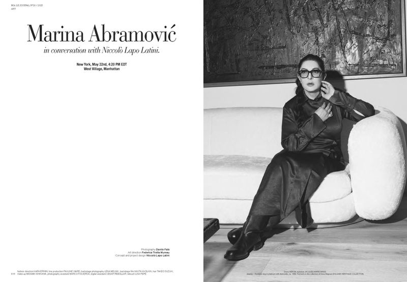 Black and white portrait of Marina Abramović, wearing dark clothing and glasses, seated on a white couch in an indoor setting.
