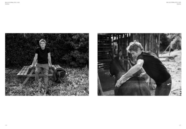 Black and white editorial shot of a man outdoors, sitting on a picnic table, alongside a man grooming a donkey by a fenced enclosure.