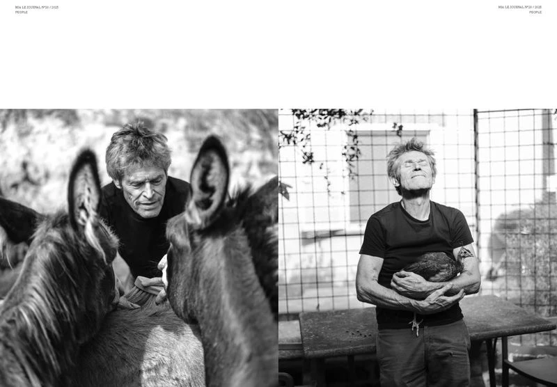 Black and white portrait of Willem Dafoe with donkeys and holding a chicken outside with a metal fence in the background.