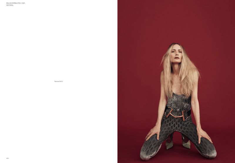 Fashion editorial shot of a woman with blonde hair wearing a silver jumpsuit kneeling against a red background in a studio setting.