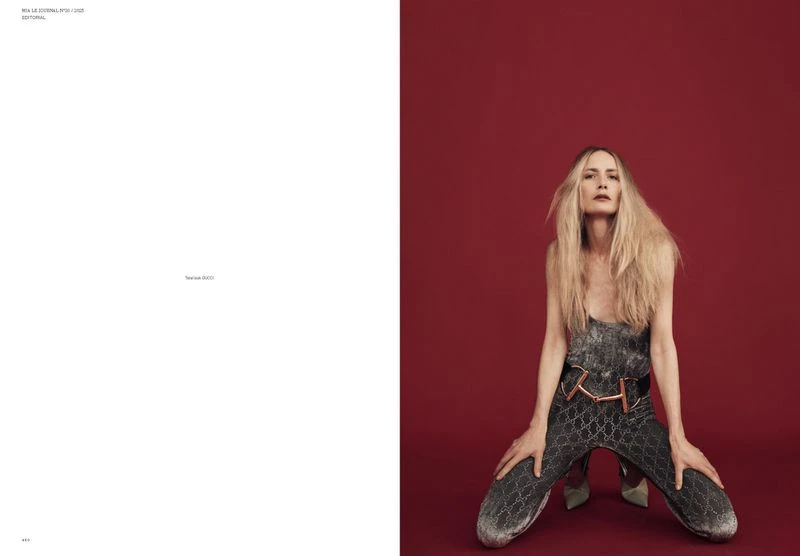 Fashion editorial shot of a woman with blonde hair wearing a silver jumpsuit kneeling against a red background in a studio setting.