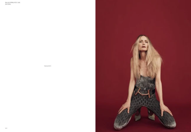 Fashion editorial shot of a woman with blonde hair wearing a silver jumpsuit kneeling against a red background in a studio setting.