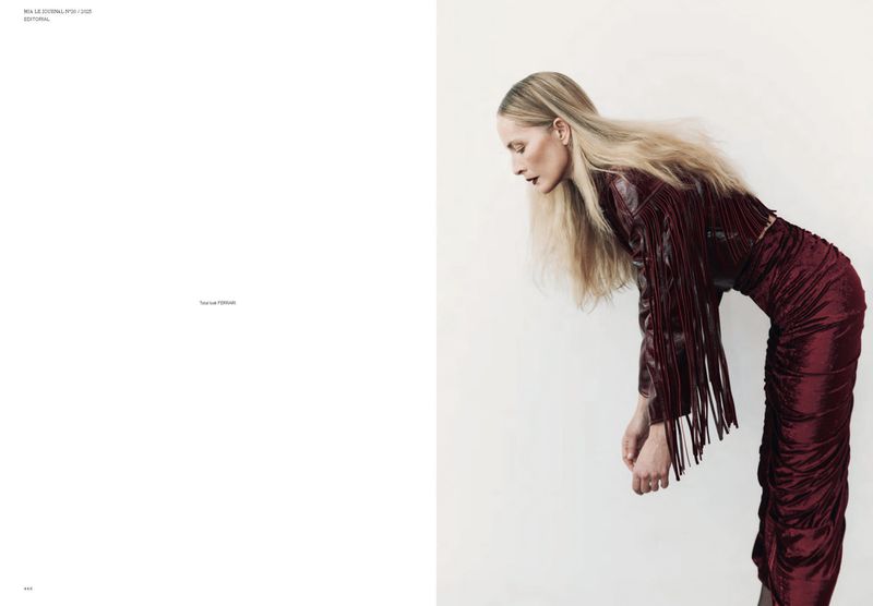Fashion editorial shot of a woman in a burgundy fringe jacket and matching ruched skirt, posed against a white wall backdrop.