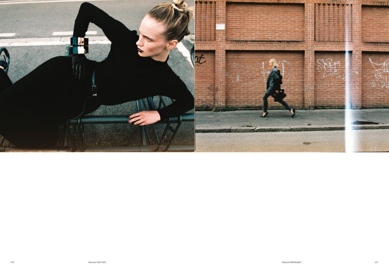 Editorial shot of a blonde woman in a black bodysuit reclining on a metal railing next to a woman walking along a brick wall.