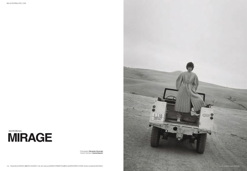 Editorial fashion photo of a woman in a long dress standing in the back of a vintage vehicle, in a hilly landscape on a cloudy day.
