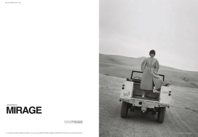 Editorial fashion photo of a woman in a long dress standing in the back of a vintage vehicle, in a hilly landscape on a cloudy day.