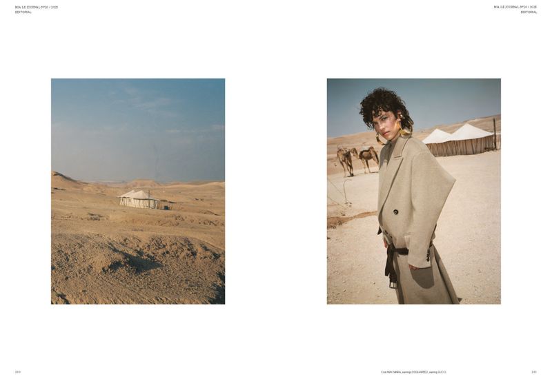 Fashion editorial photo of a woman in a tan blazer and skirt posing in the desert, with camels and tents in the background, under a blue sky.