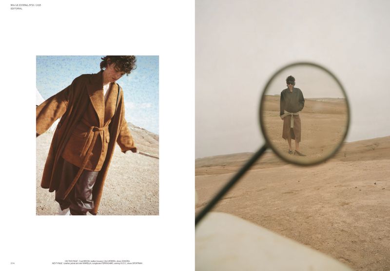 Fashion editorial featuring a young adult male model in a brown belted coat and brown leather pants posing in a desert landscape under a cloudy sky.