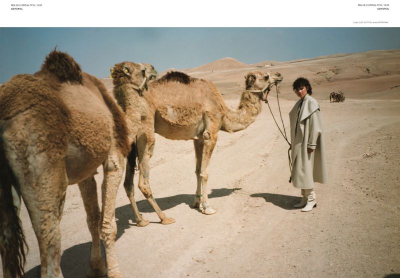 Woman in overcoat with two camels in a desert landscape, medium shot, editorial fashion photography.