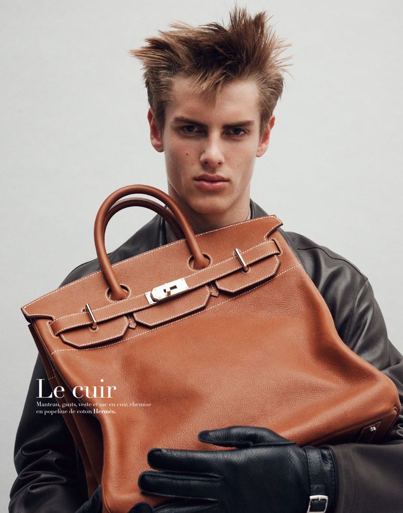 Blonde male model poses indoors with a brown leather Birkin handbag, wearing a black leather jacket and gloves against grey background.