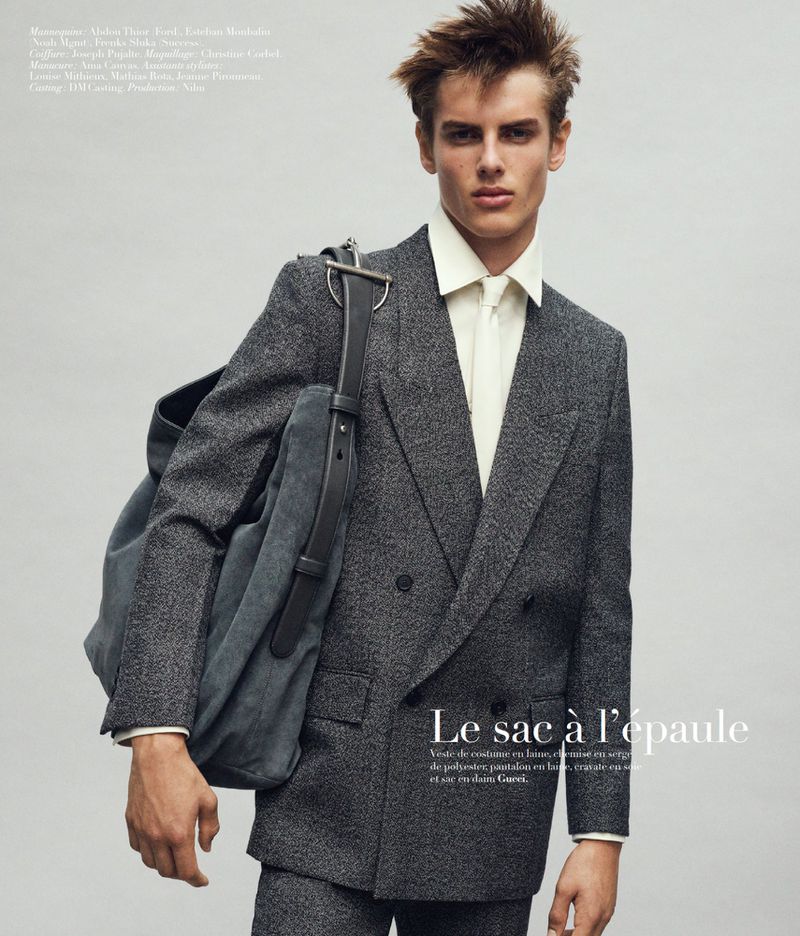 Model wearing a grey wool double-breasted suit and white shirt carrying a large shoulder bag against a neutral studio backdrop.