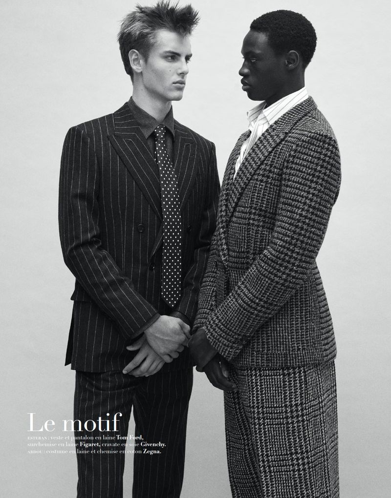 Two men in suits stand indoors. One wears a pinstripe suit with a patterned tie, while the other wears a tweed suit.