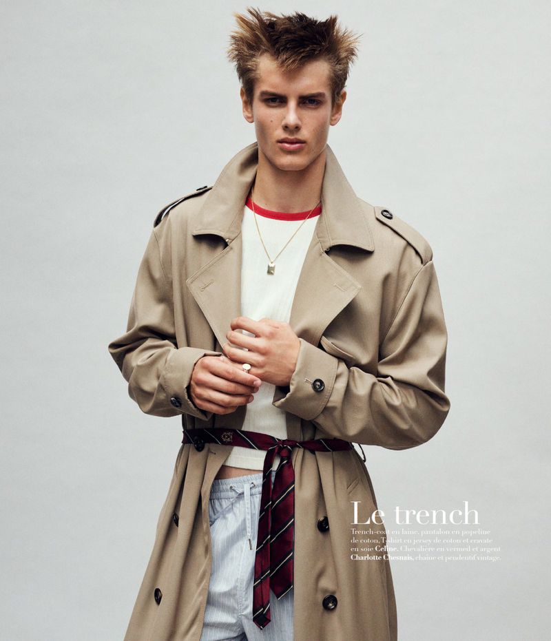 Fashion editorial shot of a young man wearing a trench coat, white shirt, and necktie belt against a neutral backdrop.