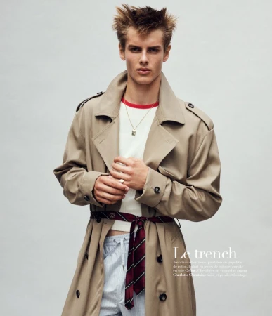 Fashion editorial shot of a young man wearing a trench coat, white shirt, and necktie belt against a neutral backdrop.