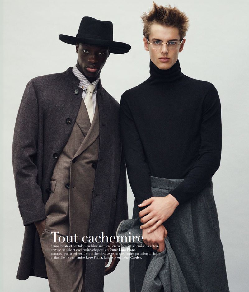 Two male fashion models wearing stylish outerwear including a hat, coat, turtle neck, glasses, suit, and holding a jacket in studio setting.