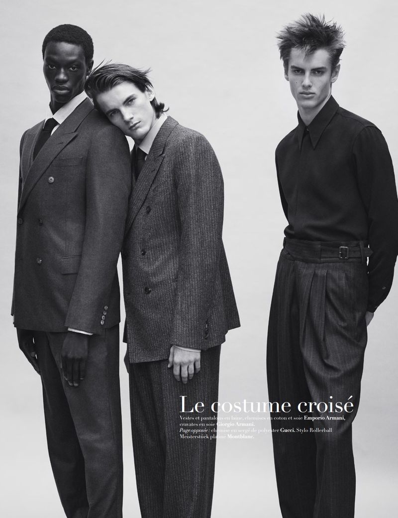 Three male fashion models pose in a black-and-white studio shot wearing suits and dress pants. The models have a neutral expression.