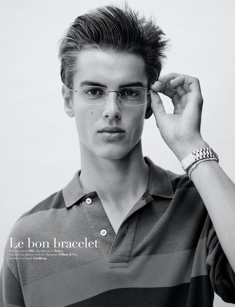 Monochrome portrait of a young adult male model wearing rimless glasses and a striped polo shirt, accessorized with a bracelet watch.