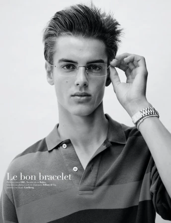 Monochrome portrait of a young adult male model wearing rimless glasses and a striped polo shirt, accessorized with a bracelet watch.