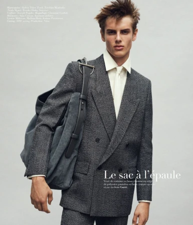 Model wearing a grey wool double-breasted suit and white shirt carrying a large shoulder bag against a neutral studio backdrop.