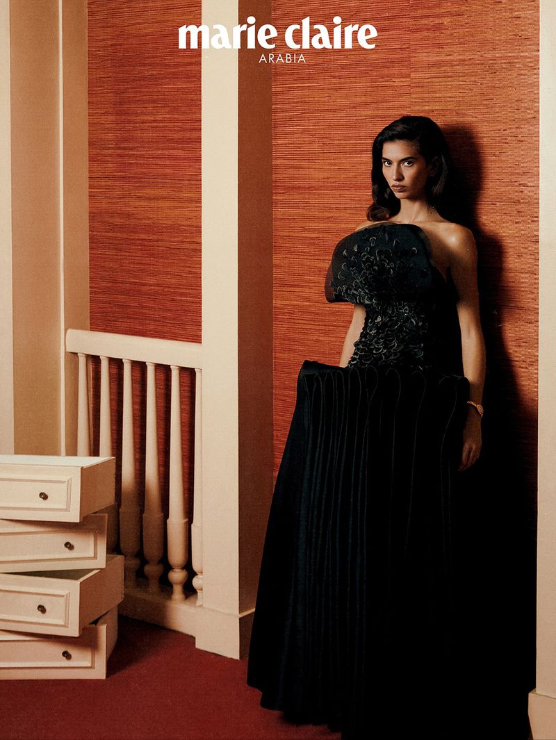 Elegant woman in a black strapless gown with floral accents posing in an indoor setting with an orange accent wall for a fashion editorial.