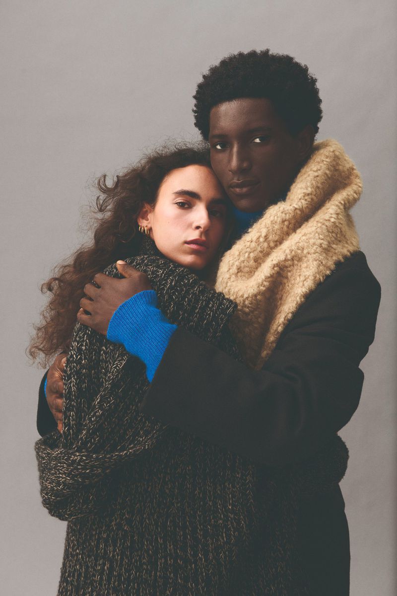 An African American man and Caucasian woman embrace in a close-up fashion shot, both wearing sweaters and a coat against a grey backdrop.