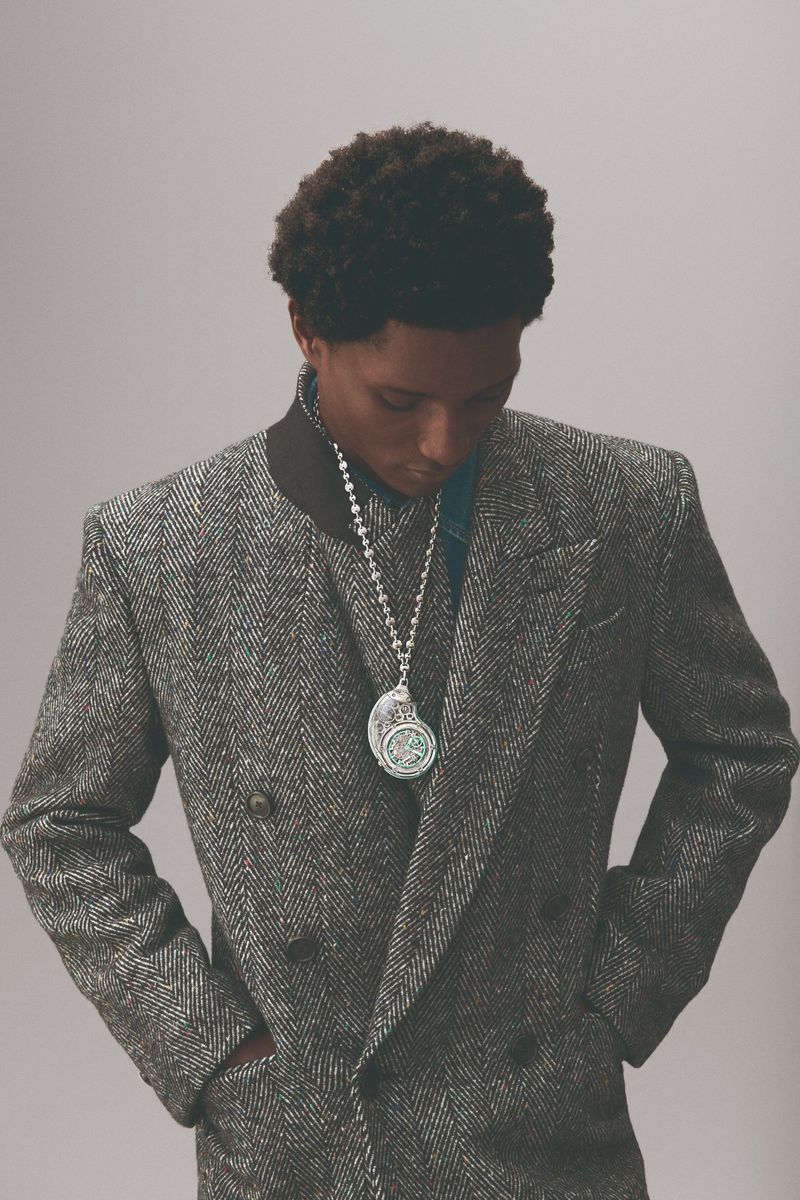 Portrait of a Black man wearing a grey tweed coat and a silver necklace with a large pendant indoors against a plain background.
