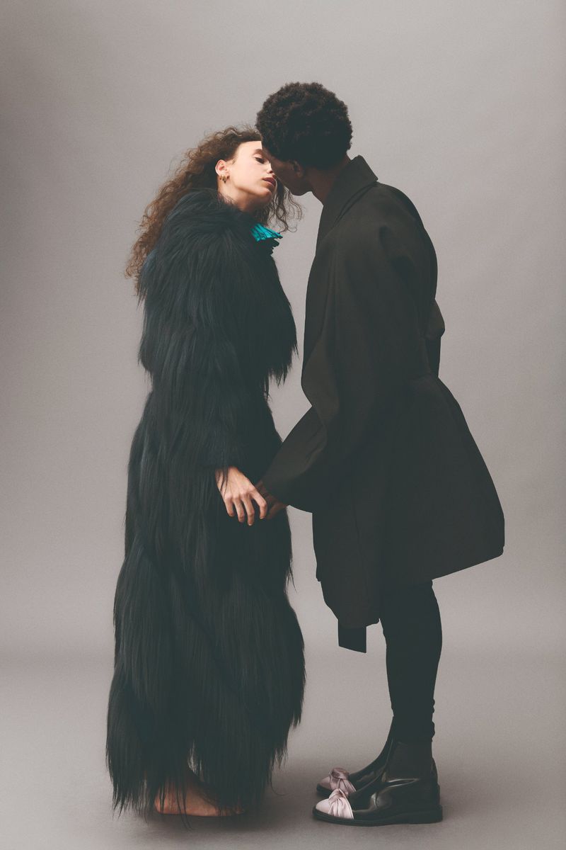 Two models in a studio setting are holding hands. One model is wearing a full length black fur coat and the other a black coat and platforms.