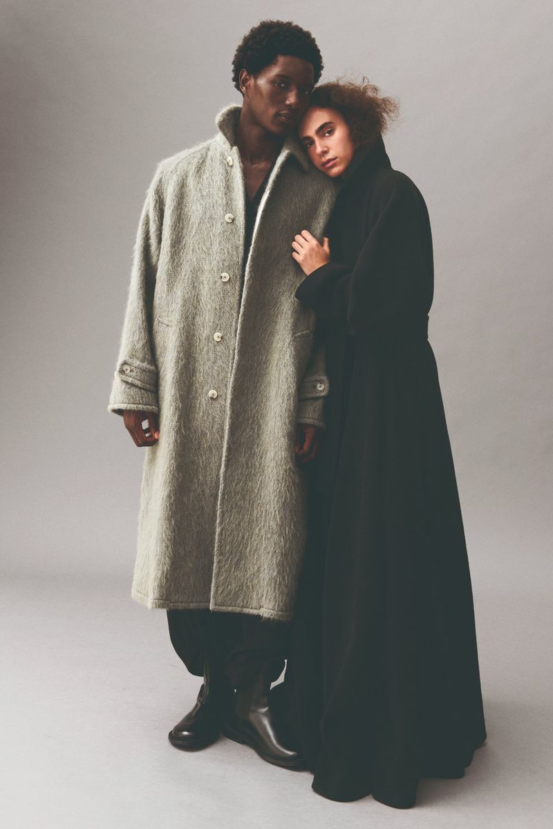 A couple posing together in stylish coats against a grey studio backdrop. The man wears a wool coat, and the woman wears a black coat.