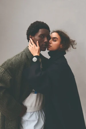 A fashion-style studio shot of a romantic couple: a Black man in a wool coat and a Caucasian woman in a black coat.