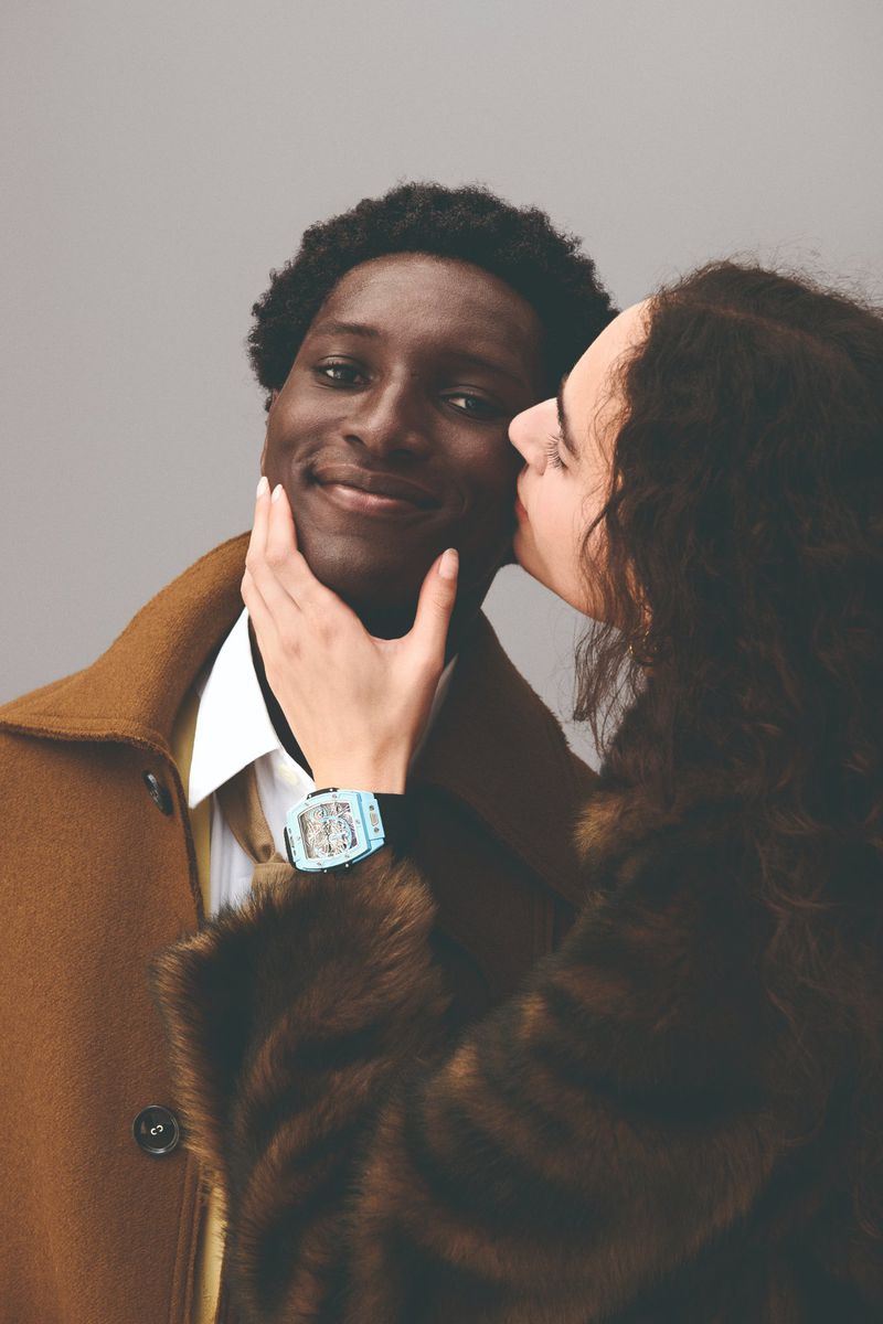 Portrait of a smiling Black man being kissed on the cheek by a white woman with curly hair, both wearing vintage-style coats.